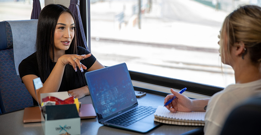 Two students are seen sitting and talking inside an Amtrak train.