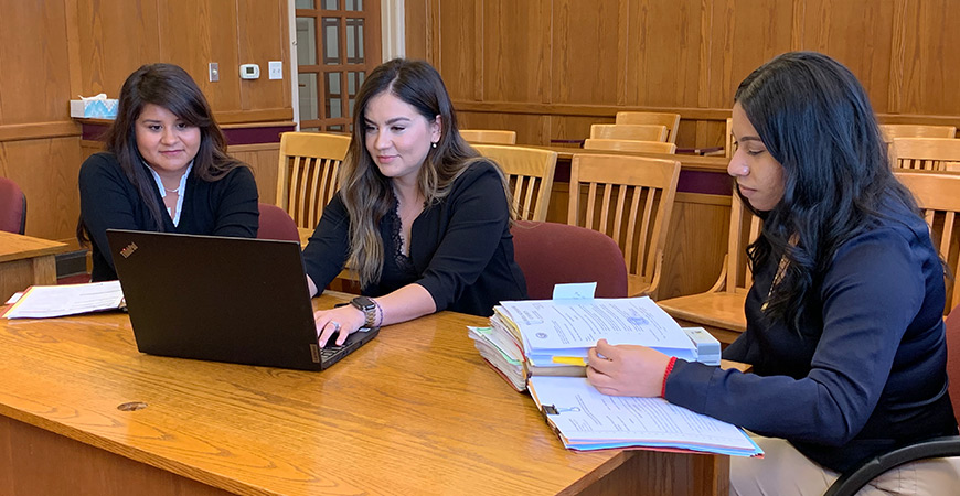 Three women sit at a desk in a mock courtroom.